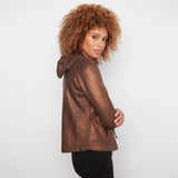 A woman with curly light brown hair is shown from the side/back wearing a metallic bronze-brown hooded sweatshirt and black pants against a plain light gray studio background.