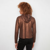 A woman with curly brown hair is shown from the back wearing a metallic bronze cropped hoodie and black jeans against a plain white background.
