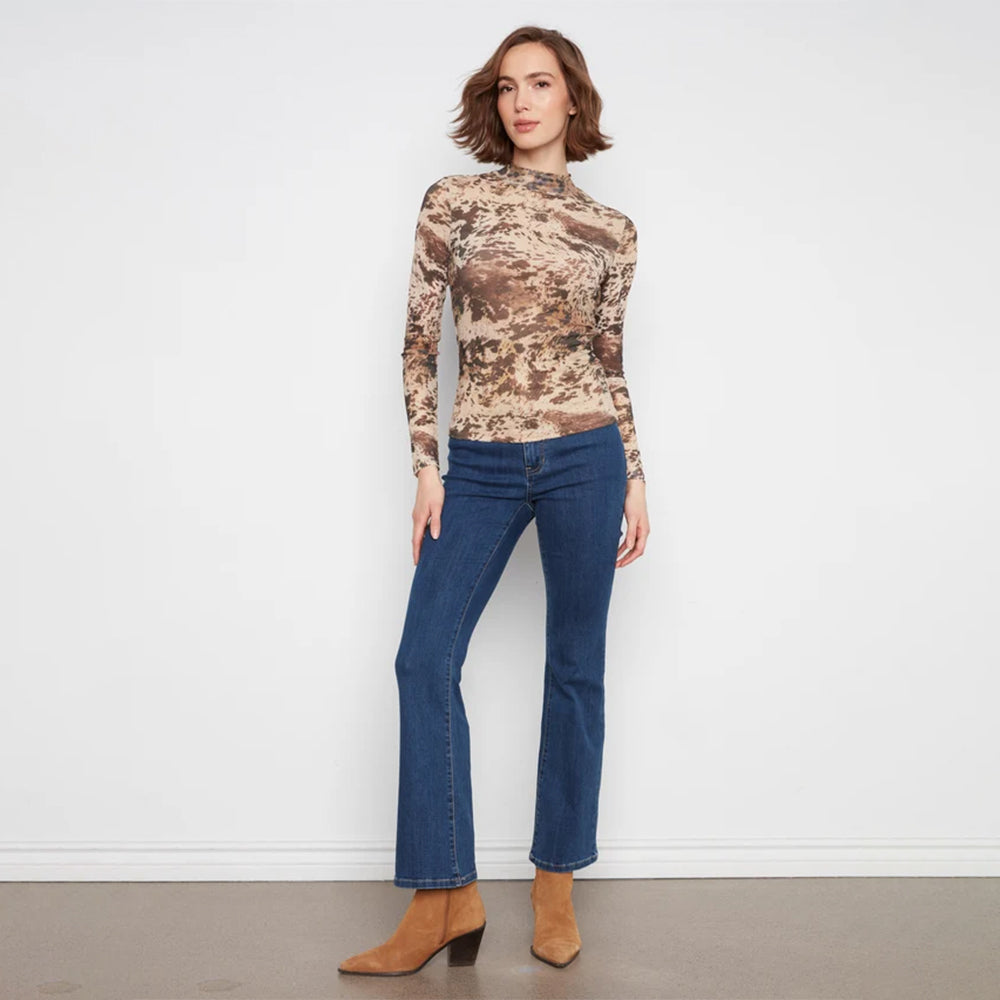 A woman stands against a white wall wearing a fitted long-sleeve mock-neck top with a brown and beige abstract print, blue jeans, and tan ankle boots.