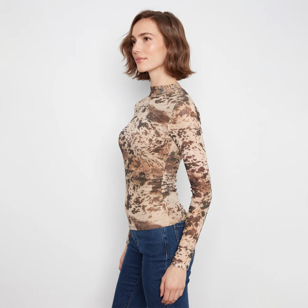 A woman wearing a fitted long-sleeve mock neck top with an abstract organic pattern in beige, brown, and tan, paired with dark blue jeans, standing against a light gray studio background.
