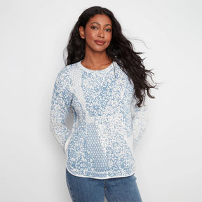 A woman with long, wavy dark hair wearing a light blue and white patterned long-sleeve knit top and blue jeans, standing against a plain white studio background.