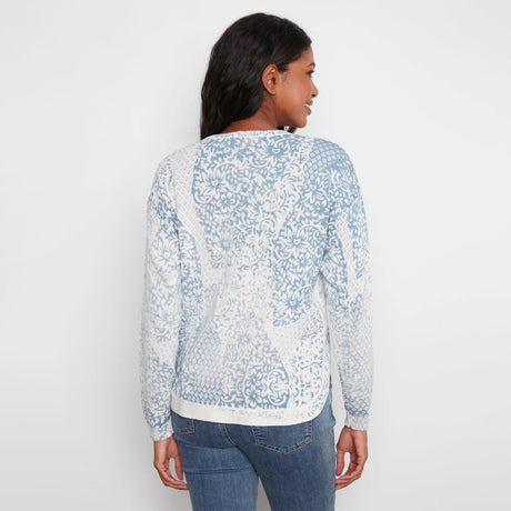 A woman viewed from the back wearing a light blue and white patterned knit sweater with blue jeans, set against a plain white studio background.