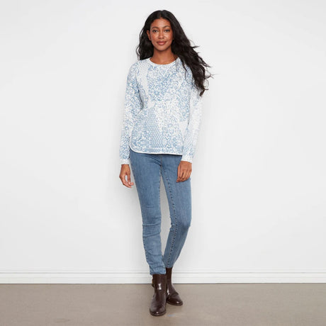 A woman modeling a light blue and white patterned long-sleeve knit top with light-wash blue jeans and dark brown ankle boots, standing against a plain white wall.