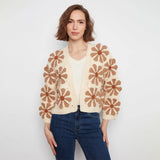 A woman modeling a cream open-front cardigan with large brown crochet flower motifs, paired with a white top and blue jeans, against a plain light gray background.