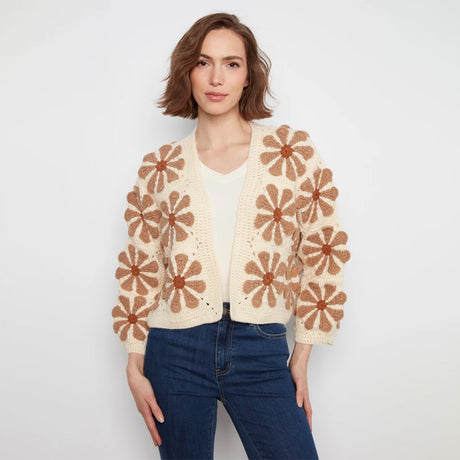 A woman modeling a cream open-front cardigan with large brown crochet flower motifs, paired with a white top and blue jeans, against a plain light gray background.
