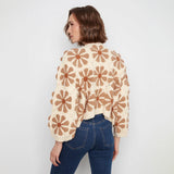 A woman shown from the back wearing a cream crochet cardigan with a brown daisy pattern, paired with blue jeans, against a plain light gray background.