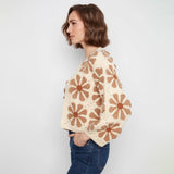 A woman wearing a cream-colored crochet sweater with brown daisy patterns, paired with blue jeans, shown from a side profile against a light gray background.