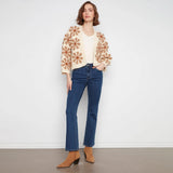 A woman stands against a plain white wall wearing a cream knit cardigan with large tan floral patterns, a cream top, blue jeans, and tan ankle boots.