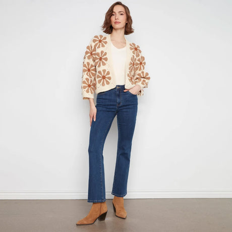 A woman stands against a plain white wall wearing a cream knit cardigan with large tan floral patterns, a cream top, blue jeans, and tan ankle boots.