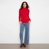 A woman stands against a plain white wall wearing a bright red ribbed mock-neck long-sleeve sweater, wide-leg blue jeans, and brown leather loafers.