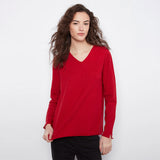 A woman with long, wavy brown hair wearing a bright red long-sleeve V-neck top and black pants, standing against a plain white studio background.