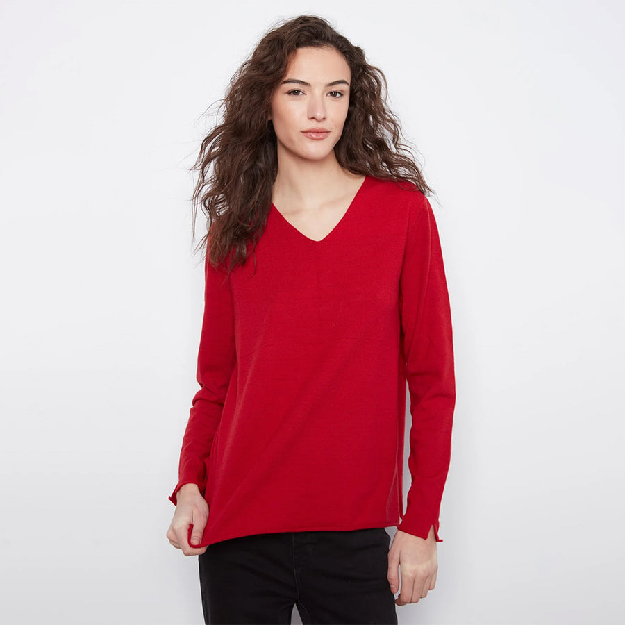 A woman with long, wavy brown hair wearing a bright red long-sleeve V-neck top and black pants, standing against a plain white studio background.