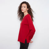 A woman with long, wavy brown hair is shown in profile wearing a bright red long-sleeve top and black pants against a plain white studio background.