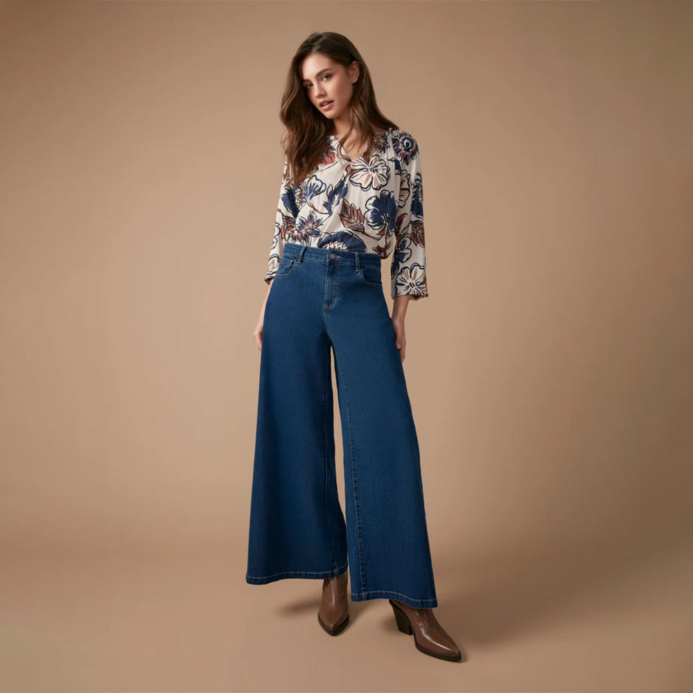 A woman modeling a pair of wide-leg blue denim jeans with a patterned blouse, standing against a solid beige studio backdrop.