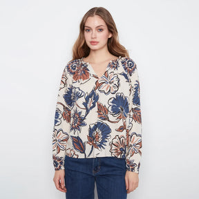 A woman modeling a long-sleeve V-neck blouse with a blue, rust, and white floral pattern on a cream background, paired with dark blue jeans, against a plain white studio background.