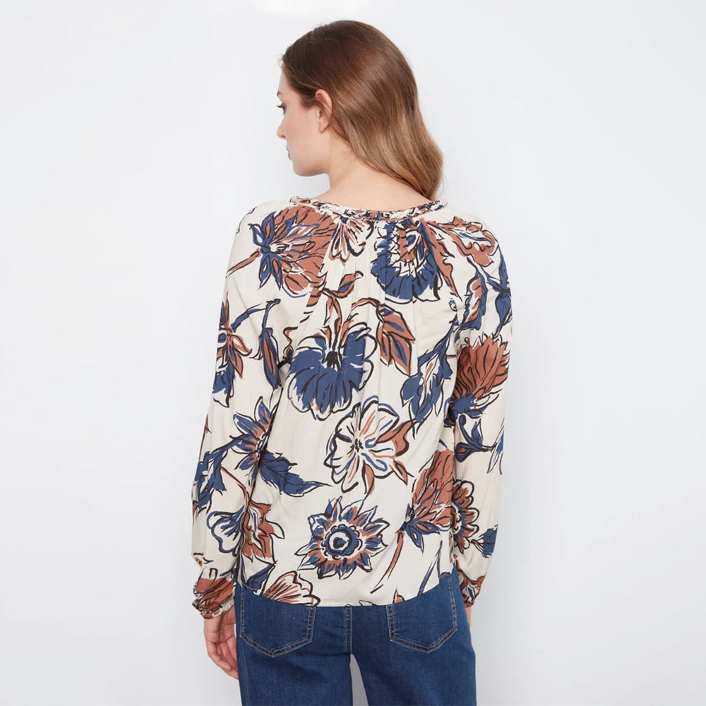 A woman viewed from the back wearing a long-sleeve cream blouse with a large blue and rust-orange floral print, paired with blue jeans, against a plain white background.