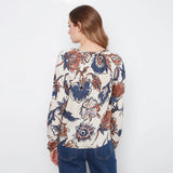 A woman viewed from the back wearing a long-sleeve cream blouse with a large blue and rust-orange floral print, paired with blue jeans, against a plain white background.
