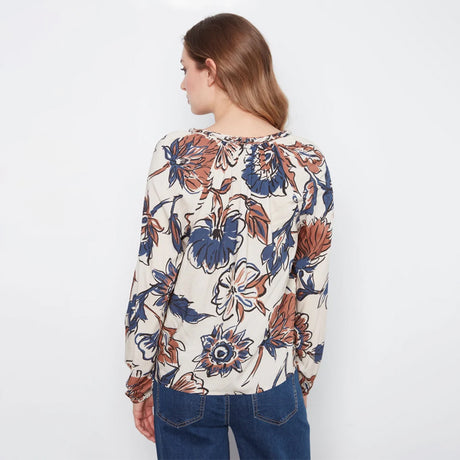 A woman viewed from the back wearing a long-sleeve cream blouse with a large blue and rust-orange floral print, paired with blue jeans, against a plain white background.