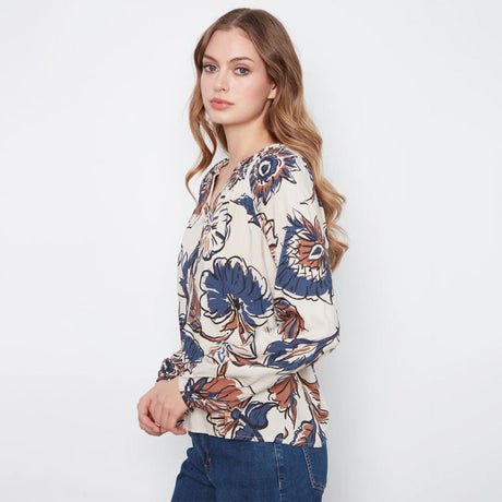A woman wearing a cream-colored long-sleeve blouse with a blue, brown, and orange floral pattern, paired with blue jeans, photographed against a plain white studio background.
