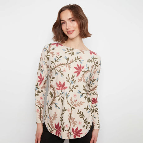 A woman wearing a long-sleeve cream-colored blouse with a colorful floral and vine pattern in pink, green, and brown, paired with black pants, set against a plain white studio background.