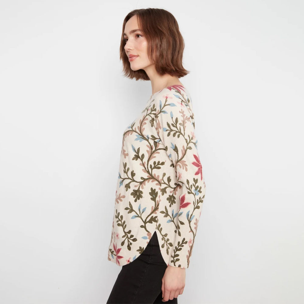 A woman with short brown hair is shown from the side, wearing a cream-colored long-sleeve top with a detailed botanical pattern in olive green, pink, and light blue, paired with black pants against a plain white background.