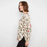 A woman with short brown hair is shown from the side, wearing a cream-colored long-sleeve top with a detailed botanical pattern in olive green, pink, and light blue, paired with black pants against a plain white background.