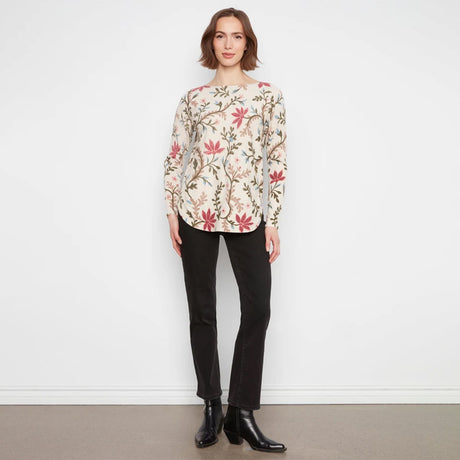 A woman stands against a white wall wearing a long-sleeve cream blouse with a red and green floral pattern, paired with black pants and black ankle boots.