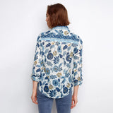 A woman viewed from the back wearing a blue and white floral patterned button-up shirt with blue jeans, set against a plain white background.