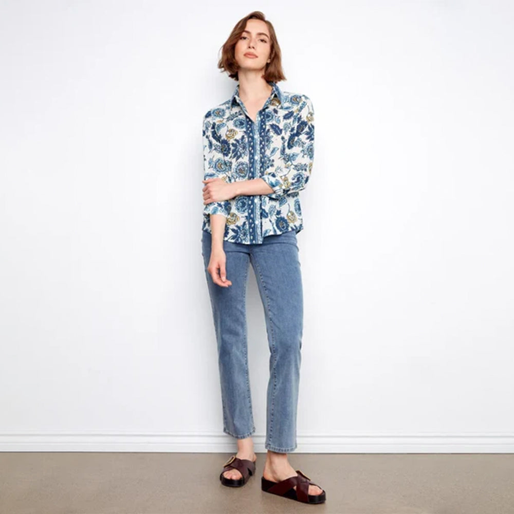 A woman stands against a white wall wearing a blue and white floral button-up shirt, light blue jeans, and dark brown slide sandals.