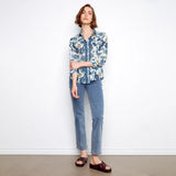A woman stands against a white wall wearing a blue and white floral button-up shirt, light blue jeans, and dark brown slide sandals.
