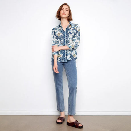A woman stands against a white wall wearing a blue and white floral button-up shirt, light blue jeans, and dark brown slide sandals.