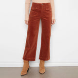 A model wearing rust-colored wide-leg corduroy pants, styled with a beige ribbed long-sleeve top and tan suede ankle boots, standing against a white wall.
