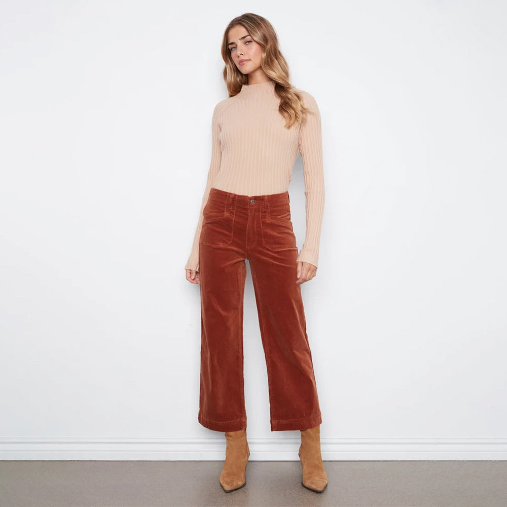 A woman stands against a plain white wall wearing a beige ribbed mock-neck long-sleeve top, rust-brown wide-leg corduroy pants, and tan ankle boots.