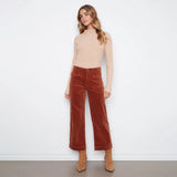 A woman stands against a plain white wall wearing a beige ribbed mock-neck long-sleeve top, rust-brown wide-leg corduroy pants, and tan ankle boots.