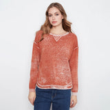 A woman modeling a rust-orange long-sleeve knit sweater with a distressed, marled texture and white contrast trim at the crew neckline, sleeve cuffs, and hem, paired with blue jeans against a plain white studio background.