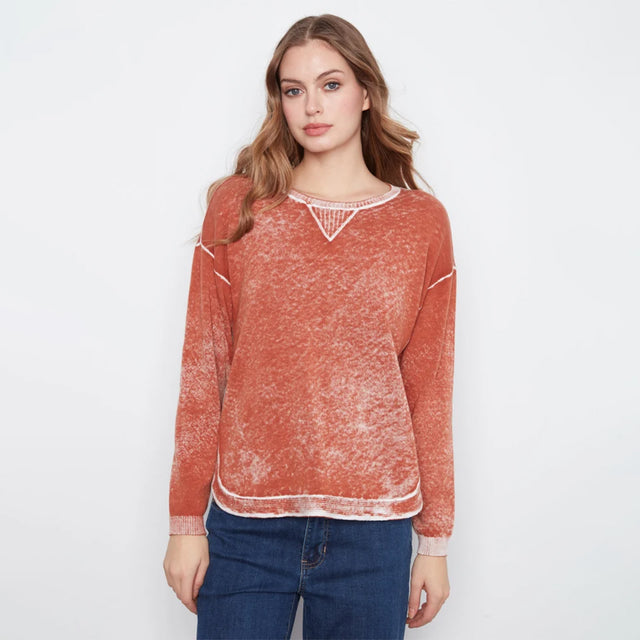 A woman modeling a rust-orange long-sleeve knit sweater with a distressed, marled texture and white contrast trim at the crew neckline, sleeve cuffs, and hem, paired with blue jeans against a plain white studio background.