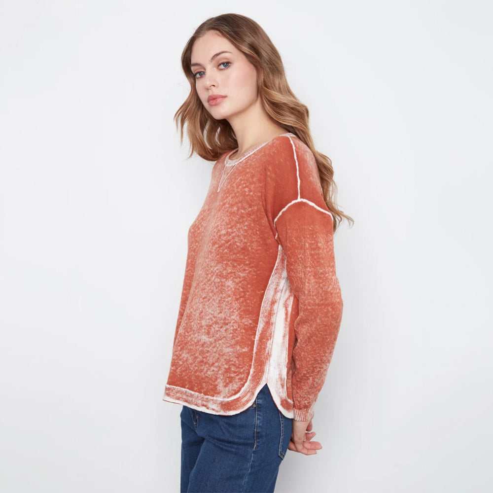 A woman modeling a rust-orange textured knit sweater with white contrast stitching, paired with blue jeans, against a plain white studio background.
