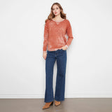 A woman stands against a white wall wearing a rust-orange textured knit sweater with white trim, dark blue wide-leg jeans, and tan ankle boots.