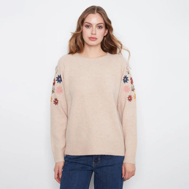 A woman modeling a beige crewneck sweater with colorful embroidered floral patterns on the sleeves, paired with blue jeans, against a plain white background.