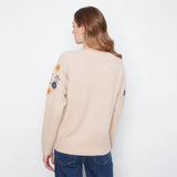 A woman shown from the back wearing a light beige knit sweater with colorful floral embroidery on the upper sleeves, paired with blue jeans, against a plain white background.