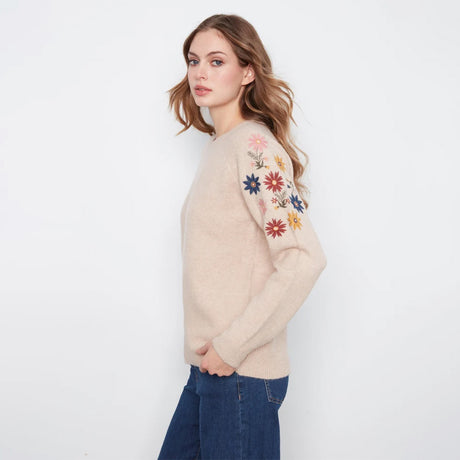 A woman modeling a beige knit sweater with colorful floral embroidery on the upper sleeve, paired with dark blue jeans, against a plain white background.
