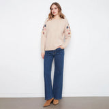 A woman stands against a white wall wearing a beige knit sweater with colorful floral embroidery on the sleeves, blue wide-leg jeans, and tan ankle boots.