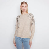 A woman modeling a beige knit sweater with leopard-print lace sleeves, paired with light blue jeans, against a plain white studio background.