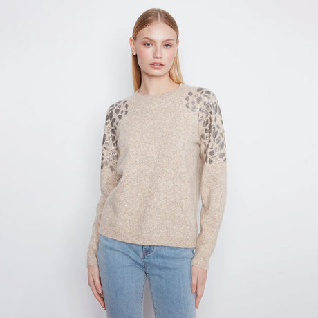 A woman modeling a beige knit sweater with leopard-print lace sleeves, paired with light blue jeans, against a plain white studio background.