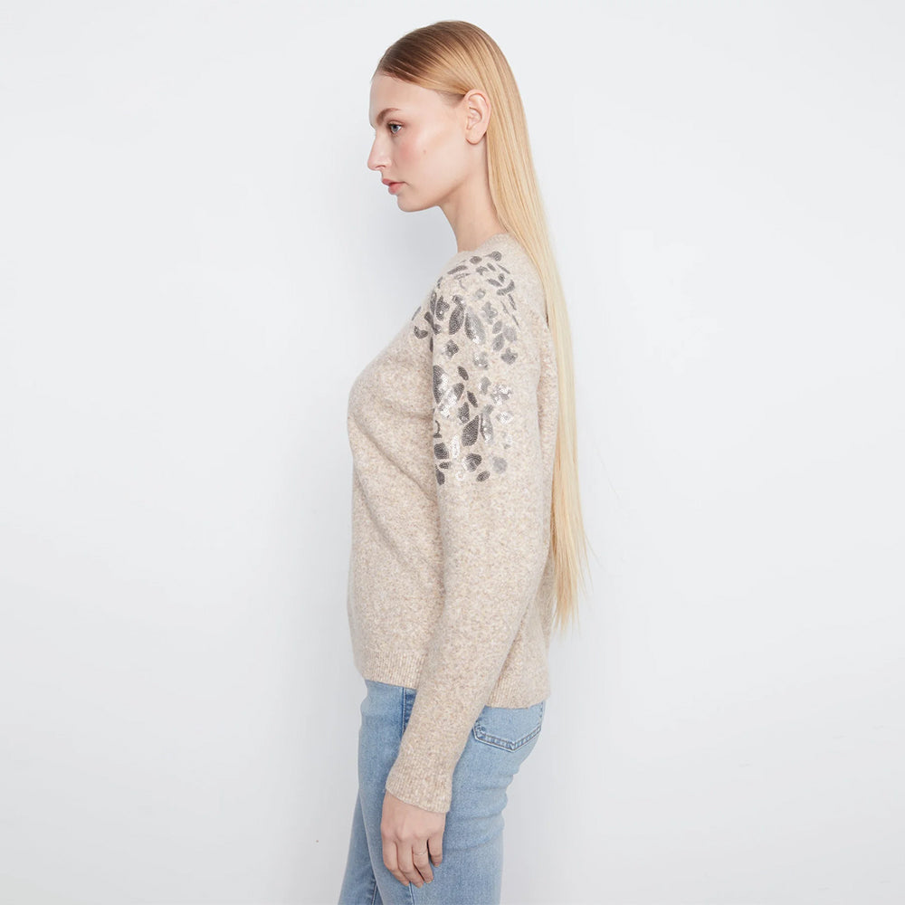 A woman in profile wearing a beige knit sweater with gray floral lace detailing on the upper sleeves, paired with light blue jeans, against a plain white studio background.