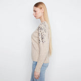 A woman in profile wearing a beige knit sweater with gray floral lace detailing on the upper sleeves, paired with light blue jeans, against a plain white studio background.