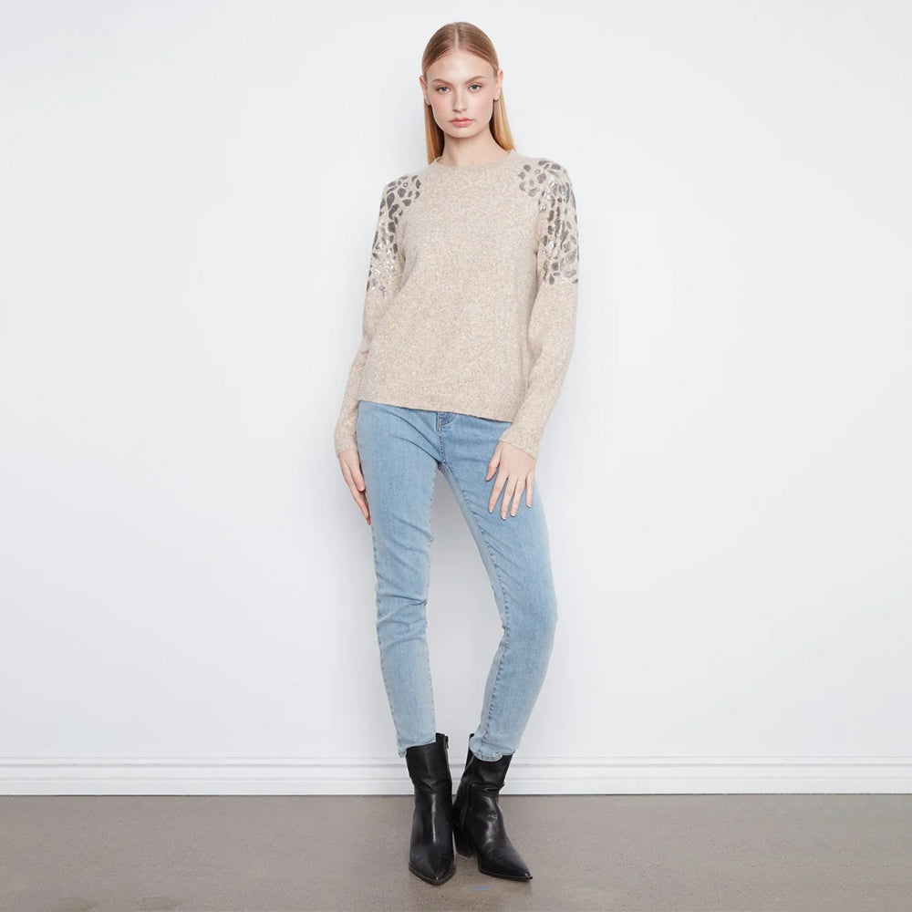 A woman modeling a beige knit sweater with textured leopard-print lace sleeves, paired with light blue skinny jeans and black ankle boots, standing against a white wall.