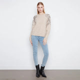 A woman modeling a beige knit sweater with textured leopard-print lace sleeves, paired with light blue skinny jeans and black ankle boots, standing against a white wall.