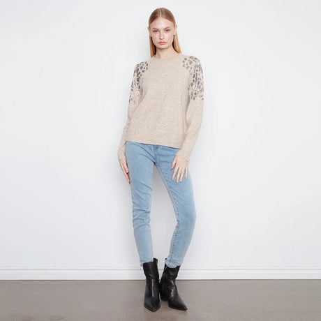 A woman modeling a beige knit sweater with textured leopard-print lace sleeves, paired with light blue skinny jeans and black ankle boots, standing against a white wall.