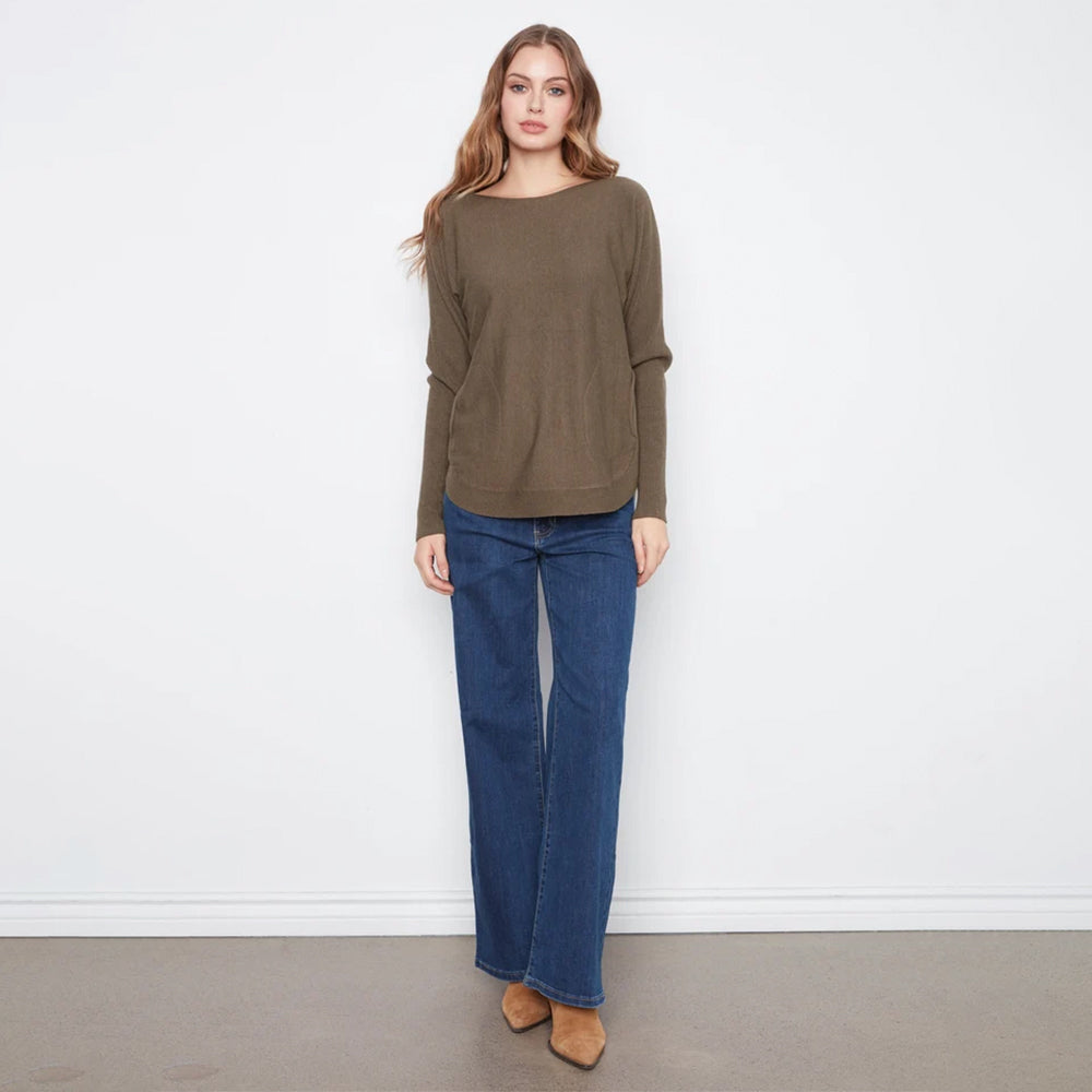 A woman modeling a loose-fitting, long-sleeve olive green sweater paired with dark blue wide-leg jeans and tan pointed-toe flats, standing against a plain white wall.